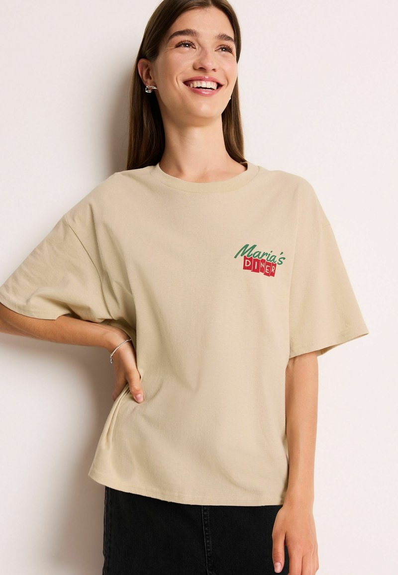 Beige katoenen T-shirt met korte mouwen, met de tekst "Maria's Diner" in rode en groene letters, losse pasvorm en een ronde halslijn.