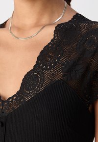Gros plan d'une femme portant un collier en argent et un haut noir avec des détails en dentelle florale au niveau de l'encolure et des épaules.