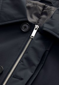 Detail čiernej bundy zobrazujúci čiastočne zapnutý strieborný zips a dva čierne gombíky pri golieri s prešitou podšívkou vo vnútri.