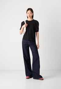 Μαύρη κοντομάνικη μπλούζα με φουσκωτά μανίκια από δαντέλα, συνδυασμένη με σκούρες μπλε φαρδιές τζιν και ροζ παπούτσια, κρατώντας μια λευκή τσάντα ώμου.