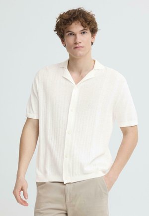 Młody mężczyzna z kręconymi włosami, ubrany w białą teksturowaną koszulę z krótkim rękawem na guziki oraz beżowe spodnie, stojący z jedną ręką w kieszeni.