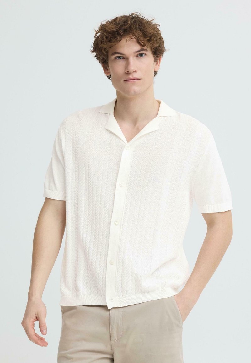 Jeune homme aux cheveux bouclés portant une chemise blanche texturée à manches courtes boutonnée et un pantalon beige, debout avec une main dans la poche.