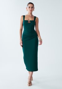 Groene aansluitende midi-jurk met vierkante halslijn, brede bandjes, voorste uitsnijding en gestructureerde stof. Gecombineerd met sandalen met bandjes in tan-kleur.