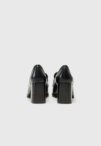 Scarpe nere in pelle con tacco alto a blocco, caratterizzate da una texture liscia e dettagli delle cuciture, viste da dietro.
