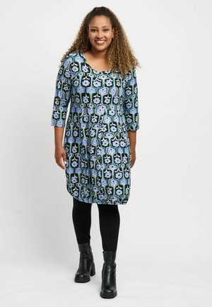 Smiling woman with curly hair wearing a black, blue, and green floral dress, black leggings, and black ankle boots standing on white background.
