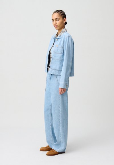 Veste en denim bleu clair avec deux poches avant, associée à un pantalon en denim bleu clair à jambe large. Accentuée par un col rayé et des chaussures beiges.