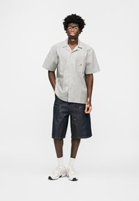 Κοντομάνικο ριγέ πουκάμισο σε λευκό και πράσινο, συνδυασμένο με σκούρα τζιν σορτς. Το μοντέλο φορά λευκά αθλητικά παπούτσια και ορατές λευκές κάλτσες.