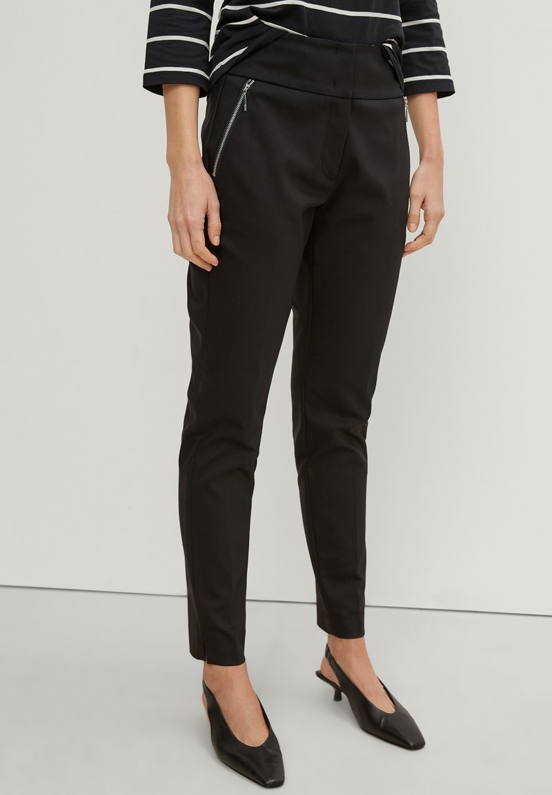 Femme portant un pantalon noir slim avec des poches avant zippées, associé à des talons slingback noirs et un haut rayé noir et blanc.