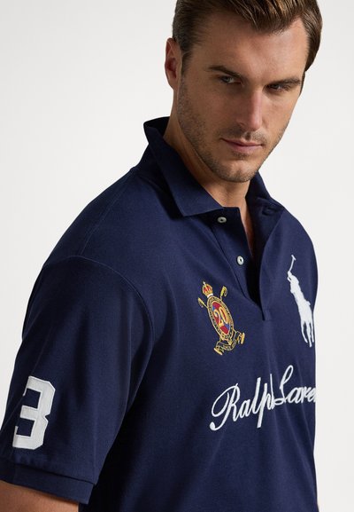 Moški, oblečen v mornarčno modro polo majico z belo izvezeno številko 3 na rokavu, grbom, igralčevim logotipom in napisom "Ralph Lauren" na prsih.