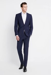 Granatowy garnitur o dopasowanym kroju, z pojedynczym zapięciem na guzik, spodniami z prostymi nogawkami i białą koszulą pod spodem. Czarne buty.