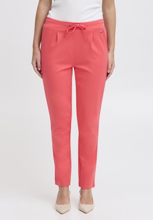 Vrouw draagt koraalroze slim-fit broek met trekkoord in de taille en beige pumps met puntige neus, staand tegen een effen achtergrond.