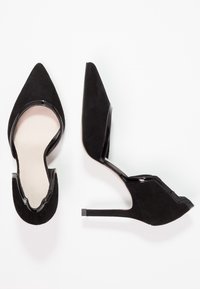 Chaussures à talons hauts noires avec un bout pointu, fabriquées en daim avec des accents en cuir verni. Design découpé sur les côtés, texture lisse.