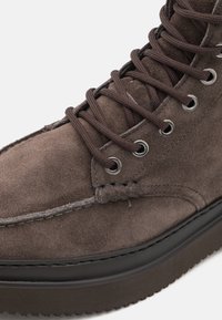 Botă ankle de culoare maro din suede, cu talpă din cauciuc negru, gauri metalice pentru șireturi și șireturi maro. Dispune de detalii de cusătură de-a lungul marginii superioare.