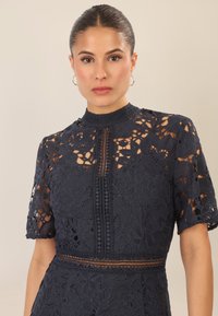Abito in pizzo blu navy con maniche corte, che presenta motivi floreali, un colletto alto e dettagli intricati di bottoni lungo la parte anteriore.