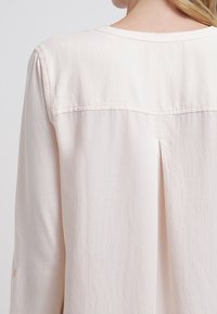 Blusa rosa chiaro caratterizzata da un tessuto morbido e leggero, scollatura tonda e una piega posteriore per una maggiore forma e movimento.