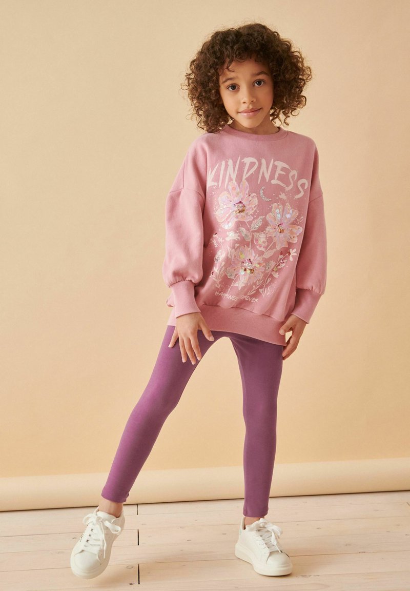 Rosa sweatshirt med blommönster och texten "KINDNESS", ihopparad med lila leggings och vita sneakers. Mjukt tyg; avslappnad passform.