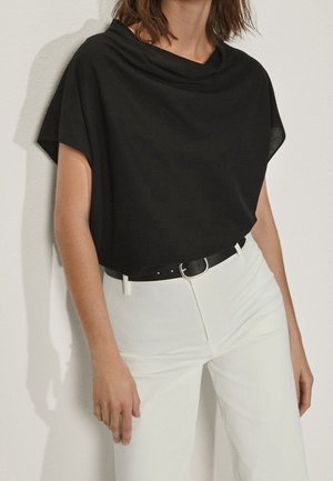 Femme portant un haut drapé noir à manches courtes, rentré dans un pantalon blanc taille haute avec une ceinture noire sur un fond uni.