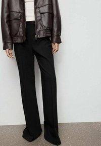 Brązowa skórzana kurtka bombowa z dwoma przednimi kieszeniami, w połączeniu z czarnymi spodniami o szerokim kroju z delikatnymi fałdami, wszystko na neutralnym tle.