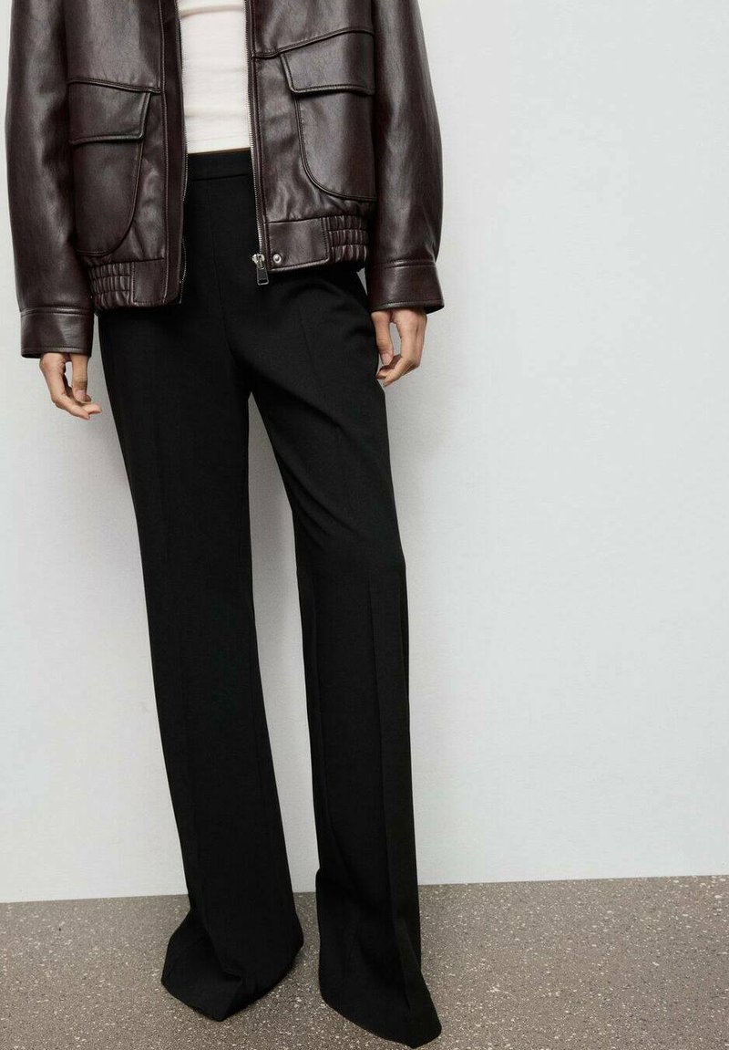 Brązowa skórzana kurtka bombowa z dwoma przednimi kieszeniami, w połączeniu z czarnymi spodniami o szerokim kroju z delikatnymi fałdami, wszystko na neutralnym tle.