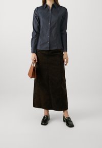Donna che indossa una camicia scura con bottoni, una lunga gonna nera, mocassini neri e tiene una piccola borsa marrone contro uno sfondo semplice.