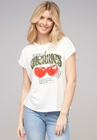 T-shirt blanc à manches courtes arborant un graphique de deux cerises rouges et un texte vert. Matière en coton doux avec une coupe décontractée.