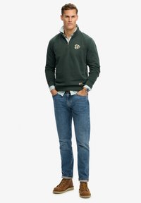 Hombre vestido con un suéter de media cremallera verde oscuro sobre una camisa a rayas, vaqueros azules, botas marrones y un reloj de pulsera, de pie con las manos en los bolsillos.