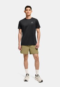 Schwarzes T-Shirt mit kurzen Ärmeln und einem Logo, kombiniert mit olivgrünen Shorts und weißen Sportsocken. Trägt hellfarbene Laufschuhe.