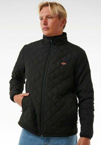 Black quilted jacket with a high collar and zip front, featuring a small brown logo on the chest and elastic cuffs. Smooth texture.
