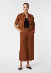 Veste cintrée brune à boutons et à manches courtes, associée à un pantalon large de la même couleur, complété par des chaussures à enfiler noires.