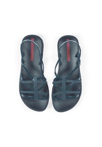 Dark teal sandals with thin, crisscross straps and textured black sole featuring a geometric pattern. Red branding on the insole.
