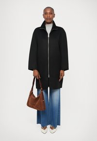 Manteau noir avec une fermeture éclair à l'avant et des manches larges, haut gris, jeans flare bleu clair, sac à main en suede marron et chaussures crème à bout pointu avec des accents.