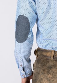 Světle modrý rukáv košile s malými černými vzory a tmavě šedou loketní nášivkou, nošený s opotřebovanými hnědými koženými kalhotami.