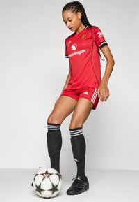 Röd fotbollskit med svarta accenter, med rund hals, logotyp och tre ränder. Svarta strumpor med vita ränder och en mönstrad fotboll.