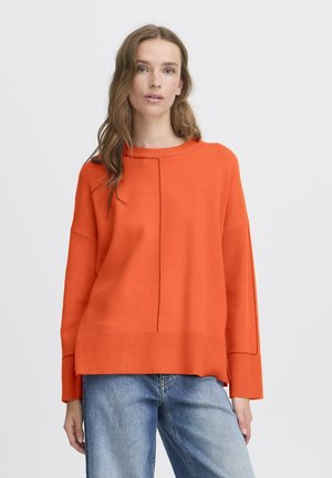 Frau mit hellbraunen Haaren, die einen übergroßen, leuchtend orangefarbenen Pullover und helle blaue Jeans trägt, vor einem einfarbigen Hintergrund.