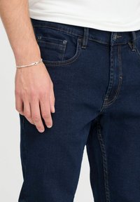 Tamno plave traperice s kontrastnim šavovima, prednjim džepovima i džepom za novčiće. Na zapešću se nosi srebrna lančić-bracelet.