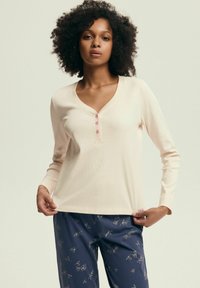 Długi rękaw, jasnoróżowa bluzka z prążkowanej tkaniny z głębokim dekoltem w kształcie litery V i trzema różowymi guzikami. Połączona z granatowymi spodniami z nadrukiem z�łotych liści.