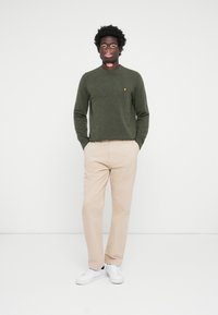 Pantalons khaki et pull vert olive à col rond avec un petit logo jaune. Le modèle porte des baskets blanches, sur un fond uni.