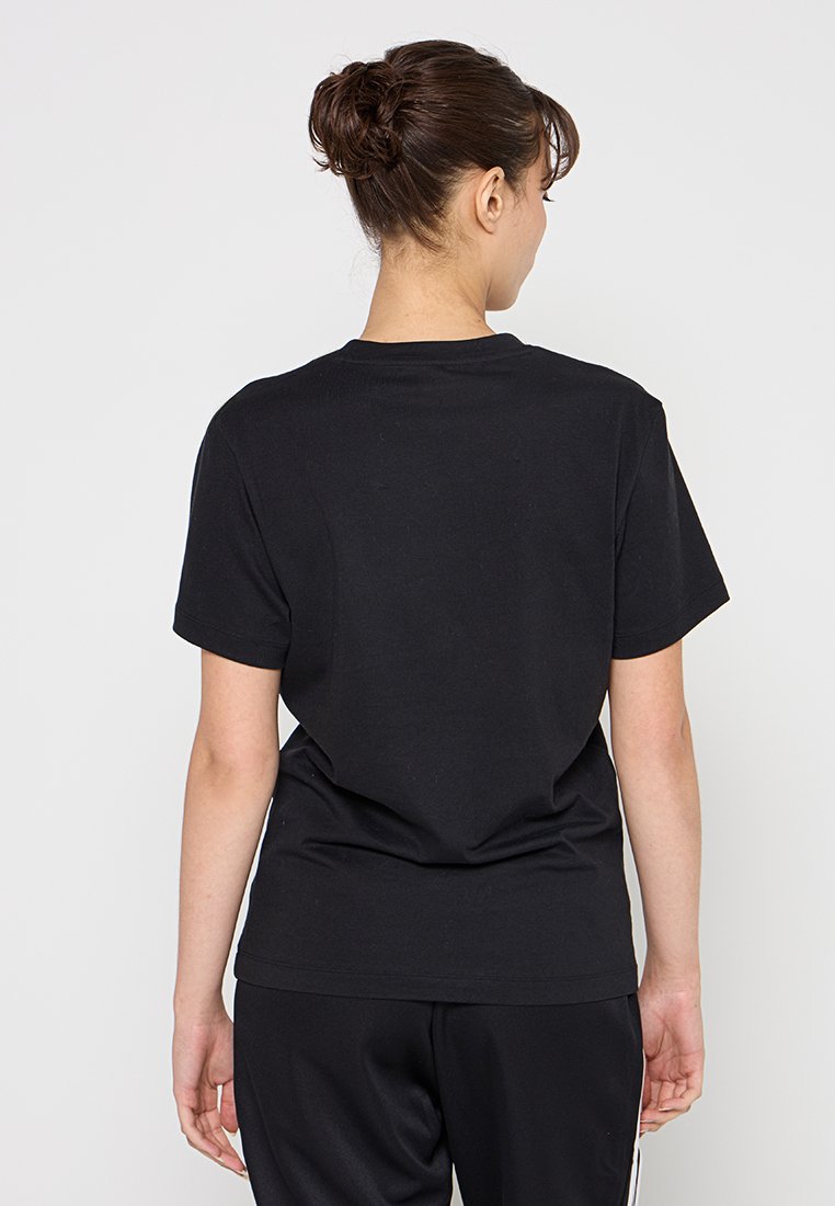 Donna con i capelli scuri raccolti in un bun, che indossa una semplice maglietta nera a maniche corte e pantaloni neri, in piedi di fronte a uno sfondo bianco uniforme.