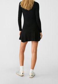 Robe noire côtelée à manches longues avec un ourlet évasé, associée à des baskets argentées brillantes et des chaussettes blanches, vue de dos.