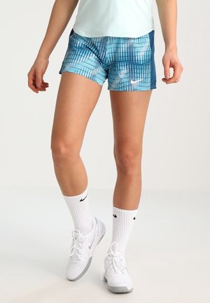 Femme portant un short de sport bleu à motifs, des chaussettes blanches montantes et des baskets blanches Nike, debout sur un fond uni clair.