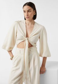 Cremfarbene Bluse mit kurzen, gepufften Ärmeln, einem tiefen V-Ausschnitt und einer vorderen Bindung. Kombiniert mit passenden hochgeschnittenen Hosen. Glattes Material.