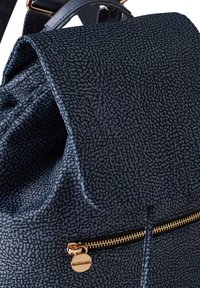 Navyblauer, strukturierten Rucksack mit Klappe, der mit einem goldenen Reißverschluss und einem kreisförmigen Metalldetail ausgestattet ist. Das Material wirkt robust und gemustert.