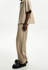 Be żowa koszula z krótkim rękawem i kołnierzykiem, zestawiona z beżowymi spodniami szerokimi w nogawkach oraz czarnymi butami o zorganizowanej formie i błyszczącej powierzchni.