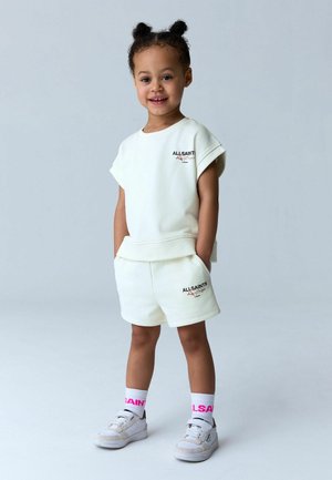 Jong kind staand, gekleed in een witte AllSaints T-shirt met korte mouwen en bijpassende korte broek, witte sokken met roze letters en witte sneakers.