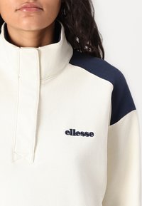 Világos krém színű, negyed cipzáros pulóver sötétkék vállrészekkel. Kiemelkedő "ellesse" logo és textúrált anyag jellemzi.