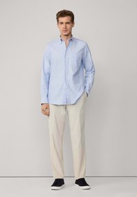 Camisa de botones azul claro con un bolsillo en el pecho, combinada con pantalones beiges y zapatillas negras. Tejido suave, diseño casual, corte estándar.