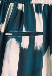 Gemusterter Stoff mit türkisfarbenen, weißen und marineblauen Streifen. Beinhaltet eine kleine, externe Tasche mit einem silbernen Etikett. Weiche Textur mit einem elastischen Bund.