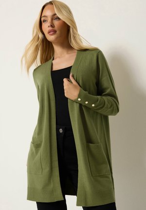 Blond meisje draagt een lange groene cardigan met zakken aan de voorkant en knoopdetails aan de manchetten over een zwart topje en zwarte broek.