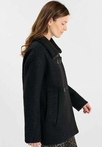 Zwarte wollen jas met een grote kraag, dubbele knopen en een zak aan de voorkant. De textuur lijkt zacht en licht pluizig.