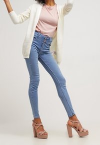 Gilet blanc léger et côtelé sur un t-shirt rose, associé à un jean skinny bleu clair taille haute et des sandales à talons blocs beiges.